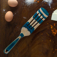 On a wooden table, there's a versatile kitchen tool: the decorative Baltique® Mykonos Collection Spurtle by Totally Bamboo. This 13" wooden spatula features colored birch and blue accents. It’s accompanied by two eggs, a block of butter, flour dust, and orange zest.