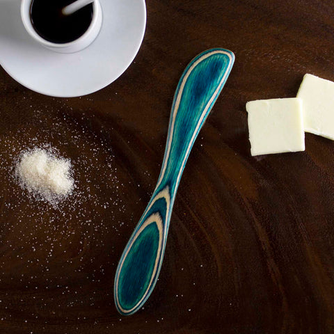 The Baltique® Mykonos Collection Spreader Knife, crafted by Totally Bamboo, features a striking blue and natural wood pattern against its brown surface. Accompanying it are two squares of butter, a small mound of salt, and a white coffee cup filled with black coffee resting on a saucer.