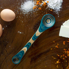 The Baltique® Mykonos Collection 2-in-1 Measuring Spoon from Totally Bamboo, crafted from colored birch with "1/4 tsp" engraved on it, is nestled among two eggs, a sprinkle of flour, shredded orange zest, and a block of butter on a wooden surface.