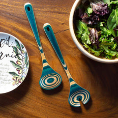 The Baltique® Mykonos Collection Salad Serving Set by Totally Bamboo, measuring 11-3/4 inches, is crafted from colored birch and features a vibrant, swirling pattern. It rests beside a bowl of fresh mixed greens and a decorative plate with olive illustrations on a wooden surface.