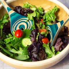 A wooden bowl filled with fresh salad, showcasing leafy greens, cherry tomatoes, and cucumber slices. The Baltique® Mykonos Collection Salad Serving Set from Totally Bamboo, featuring servers crafted from colored birch with a vibrant swirling pattern, elegantly rests on top.