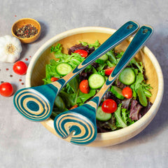 A wooden bowl brimming with fresh salad, including cherry tomatoes, cucumbers, and mixed greens, is elegantly adorned with two pieces from the Totally Bamboo Baltique® Mykonos Collection Salad Serving Set, measuring 11-3/4 inches. These pieces feature beautiful colored birch in blue and natural patterns. Nearby on a gray surface lie garlic cloves and a small bowl of peppercorns.