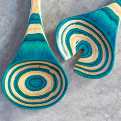 From the Baltique® Mykonos Collection by Totally Bamboo, this 11-3/4" salad serving set includes two wooden utensils adorned with a lively pattern in shades of teal, beige, and light brown. One utensil features a circular swirl design while the other displays a fork-like cutout, both set against a textured gray backdrop.