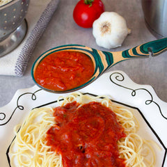 A plate of spaghetti draped in rich tomato sauce sits below a Baltique® Mykonos Collection Notched Cooking Spoon by Totally Bamboo, poised to add more deliciousness. A colorful Mykonos Collection colander enhances the scene's charm, accompanied by a fresh tomato and garlic on a gray surface, whetting the appetite for culinary delight.