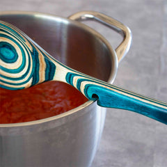 The Baltique® Mykonos Collection Notched Cooking Spoon by Totally Bamboo is crafted from Colored Birch, featuring blue and green swirls. It rests perfectly on the rim of a stainless steel pot filled with tomato sauce, thanks to its notched handle for added convenience.