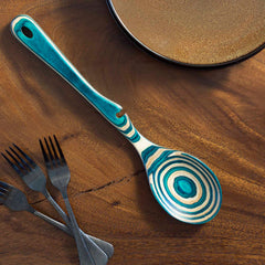 A Baltique® Mykonos Collection Notched Cooking Spoon from Totally Bamboo, featuring a unique swirl of blue and beige, lies on a wooden table. Nearby, three decorative forks with geometric patterns rest next to a subtly textured brown ceramic plate.