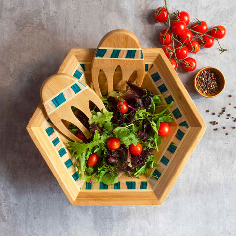 The Totally Bamboo Baltique® Mykonos Collection Salad Bowl & Salad Hands Set presents a fresh green salad adorned with cherry tomatoes. Nearby, a small dish of mixed peppercorns and a vine of cherry tomatoes can be found, while two elegant wooden salad hands rest within the bamboo bowl on a stone-textured surface.