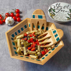 A colorful pasta salad featuring cherry tomatoes, olives, and peppers is served in the Baltique® Mykonos Collection Salad Bowl from Totally Bamboo. Accompanying the dish are the bamboo salad hands for serving. On the side, fresh cherry tomatoes and garlic complement a nearby decorative plate.