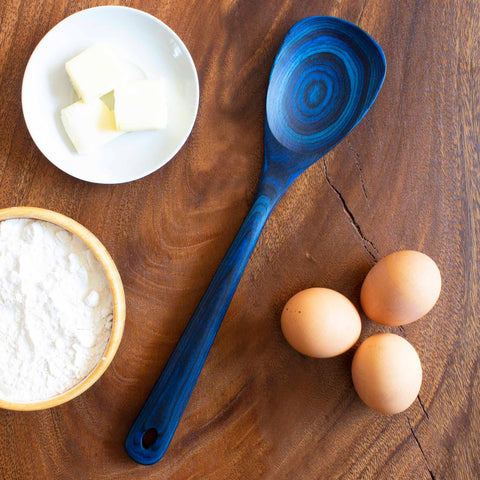 A wooden table with a Totally Bamboo Baltique® Malta Collection Cooking Spoon, 12-1/2", three brown eggs, a small bowl of flour, and a small plate with butter cubes.