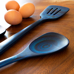 A close-up of wooden kitchen utensils showcases the colorful swirl design of a Totatly Bamboo Baltique® Malta Collection Cooking Spoon, 12-1/2", accompanied by a slotted spatula on a wooden surface. Four brown eggs can be seen in the background.