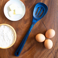 A Baltique® Malta Collection Slotted Spoon, 12-1/2" by Totally Bamboo, crafted from colored birch in a vibrant blue hue, is elegantly placed on a wooden table. It's accompanied by a white plate of butter cubes, a bowl of flour, and three eggs—a perfect setting for anyone who appreciates the art of using quality kitchen utensils.