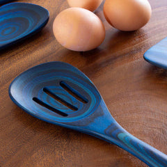 A blue Baltique® Malta Collection Slotted Spoon, 12-1/2" by Totally Bamboo rests on a wooden surface beside three brown eggs. The kitchen utensil showcases swirling patterns, and parts of blue-colored birch plates are also visible in the background.