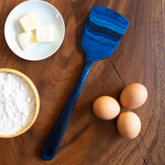 A vibrant Totally Bamboo Baltique® Malta Collection Spatula, measuring 12-1/2 inches, rests on a wooden surface, surrounded by a plate of butter, a bowl of flour, and three brown eggs.