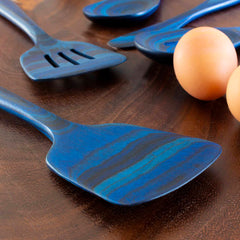 A Baltique® Malta Collection Spatula, 12-1/2" from Totally Bamboo, featuring a blue swirled pattern crafted from colored birch, lies on a dark wooden surface alongside other similarly designed kitchen utensils. Nearby are three brown eggs.