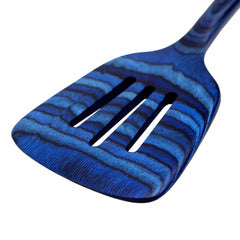 A close-up of the Baltique® Malta Collection Slotted Spatula, 12-1/2" from Totally Bamboo reveals its vibrant charm. This blue, striped spatula features a textured handle and is crafted from colored birch, showcasing an intricate design that highlights its unique and decorative appearance.