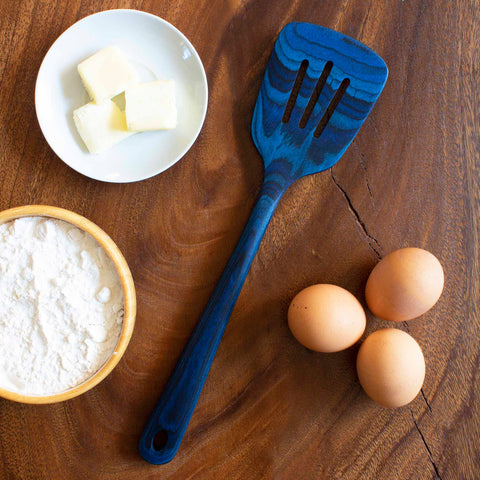 A blue slotted spatula from the Baltique® Malta Collection by Totally Bamboo rests on a wooden surface next to three brown eggs, a small round white dish with butter cubes, and a round wooden bowl filled with flour.