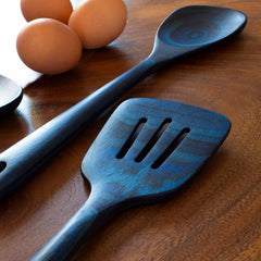 A close-up view of three brown eggs next to a Totally Bamboo Baltique® Malta Collection Slotted Spatula, 12-1/2", and a spoon on a wooden surface. The spatula boasts three slots and showcases a blue-tinged finish reminiscent of colored birch.