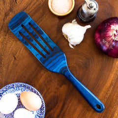 A Baltique® Malta Collection Spurtle, 13" by Totally Bamboo, with a blue finish, rests on a wooden table. It's surrounded by a garlic bulb, red onion, small bowl of salt, pepper grinder, and three eggs in a decorative bowl—perfect companions for your non-stick cookware adventures.