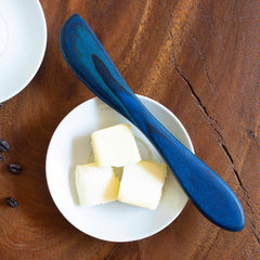 A spreader knife from the Baltique® Malta Collection by Totally Bamboo, featuring a blue design and crafted from colored birch, rests elegantly on three cubes of butter in a white dish on a wooden surface. Part of a white plate can be seen to the left.