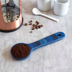 A Totally Bamboo Baltique® Malta Collection Coffee Scoop, in blue, is filled with ground coffee on a gray surface. Nearby, scattered coffee beans, a French press, white mugs, and an extra spoon complete the ideal setting for a coffee lover.