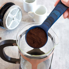 A hand holds a Totally Bamboo Baltique® Malta Collection Coffee Scoop, filled with ground coffee, over a French press. The press rests on a gray surface, accompanied by its lid and two white mugs in the background.