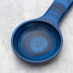 A close-up of the Totally Bamboo Baltique® Malta Collection Coffee Scoop, featuring a deep blue, round wooden design resting on a light gray surface. The texture and grain are visible, showcasing circular patterns in varying shades of blue.