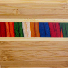 This piece of art showcases a textured design incorporating multicolored fibers that evoke the Baltique® Marrakesh Collection by Totally Bamboo, set within light wood panels displaying natural grain patterns. The fibers exhibit vivid hues of red, green, cream, blue, and orange.