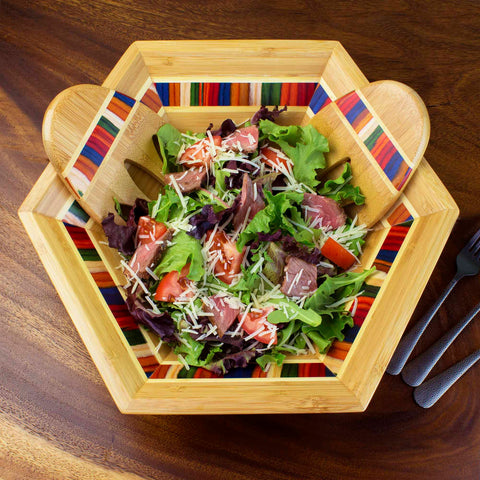 The Baltique® Marrakesh Collection Salad Bowl & Salad Hands Set by Totally Bamboo, featuring vibrant striped edges, elegantly cradles a fresh salad. The bowl is filled with mixed greens, sliced tomatoes, and shredded cheese, which are stirred using the wooden utensils. This stylish bamboo salad bowl rests on a wooden table beside two forks.