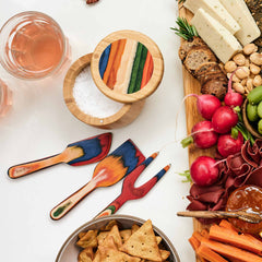 A vibrant wooden kitchen utensil set from the Baltique® Marrakesh Collection showcases rainbow colors beside a charcuterie board brimming with colorful vegetables, cheeses, and meats. An 8-ounce capacity salt cellar from Totally Bamboo serves as a charming addition with a glass of rosé and a bowl of crackers completing the scene.