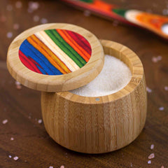 An 8-ounce Baltique® Marrakesh Collection Salt Cellar by Totally Bamboo features a colorful striped lid and is open to reveal white granulated sugar inside. It sits on a wooden surface with pink salt grains scattered around, showcasing its charming design.