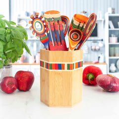 The Totally Bamboo Baltique® Marrakesh Collection Kitchen Utensil Holder, crafted from vibrant Moso bamboo, stands on the countertop holding colorful striped tools. Fresh apples, a red bell pepper, and a basil plant surround this utensil holder, set against a kitchen background of blurred shelves and appliances.