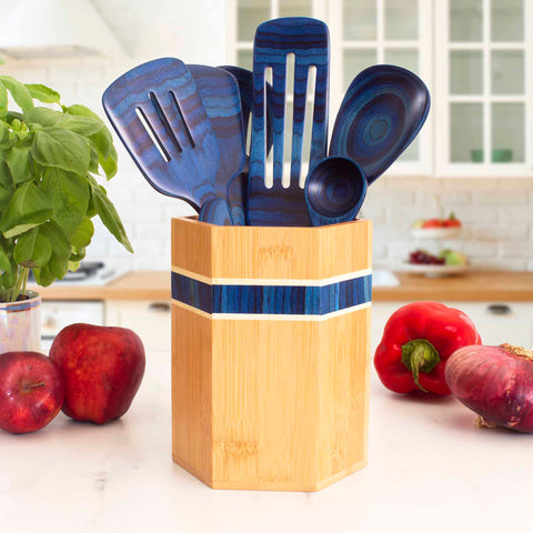 A Totally Bamboo Baltique® Malta Collection Kitchen Utensil Holder filled with blue kitchen utensils stands on a white countertop. Fresh basil, red apples, a bell pepper, and a partially visible eggplant are nearby in this bright, modern kitchen.