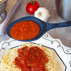 A serving of spaghetti on a decorative plate topped with tomato sauce is highlighted by a Totally Bamboo Baltique® Malta Collection Grand Serving Spoon, 14", expertly crafted from colored birch, which holds more sauce above the dish. In the background, a tomato, garlic, and a colander are visible on the counter.