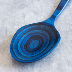 This 14" Grand Serving Spoon from Totally Bamboo's Baltique® Malta Collection features black circular patterns on a blue wooden surface, expertly crafted from colored birch and set against a gray textured background.