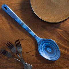 A Baltique® Malta Collection Grand Serving Spoon by Totally Bamboo, featuring a blue swirled pattern and measuring 14 inches, is elegantly placed on a wooden surface next to a circular brown ceramic plate. Nearby, three black forks with intricate designs add to the rustic and artistic composition, showcasing rich textures and colors.