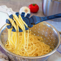 A serving of cooked spaghetti is lifted from a colander with a Baltique® Malta Collection Spaghetti Spoon, 12-1/2" by Totally Bamboo. In the soft-focus background, a garlic bulb, a tomato, and non-stick cookware rest on the kitchen countertop.