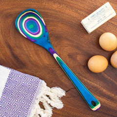 Part of the Baltique® Mumbai Collection by Totally Bamboo, this 12-1/2" multicolored cooking spoon showcases a vibrant pattern. It rests elegantly on a wooden surface alongside a bar of butter, three eggs, and a decorative purple and white towel. Crafted from colored birch, it is both functional and stylish for any kitchen setting.