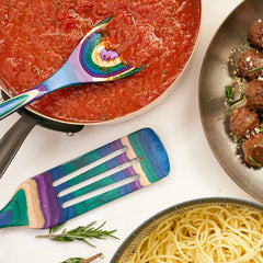 A vibrant wooden spoon and the Baltique® Mumbai Collection Spurtle, 13" by Totally Bamboo are placed next to a pan of tomato sauce and a plate of meatballs topped with rosemary and cheese. Nearby is a bowl of cooked spaghetti, each colorful kitchen tool safe for non-stick surfaces, all displayed on a light surface.