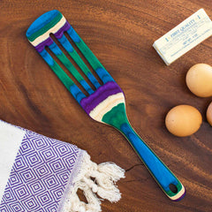 A colorful 13" Baltique® Mumbai Collection Spurtle by Totally Bamboo, designed with a non-stick safe wooden finish, elegantly sits on a wooden surface. Nearby, three brown eggs, a wrapped stick of butter, and a fringed purple and white towel with a geometric pattern accentuate this vibrant kitchen tool.