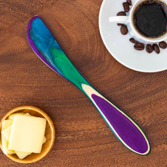 A striking spreader knife from Totally Bamboo's Baltique® Mumbai Collection, featuring a vibrant multicolored birch handle in shades of blue, green, and purple, rests on a wooden surface next to a small plate with butter and a cup of black coffee adorned with scattered beans.