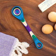 A colorful Totally Bamboo Baltique® Mumbai Collection 2-in-1 Measuring Spoon with rainbow rings is placed on a wooden surface next to a stick of butter, three eggs, and a purple geometric-patterned cloth adorned with tassels, capturing India peacock inspiration through its vibrant hues.