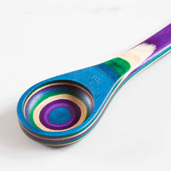 A vibrant wooden spoon from Totally Bamboo's Baltique® Mumbai Collection, showcasing a design inspired by the Indian peacock with layered stripes of blue, purple, green, and natural wood set against a white backdrop. The handle of the 2-in-1 Measuring Spoon is conveniently marked with "1 TBS.