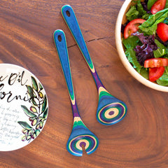 A pair of vibrant salad servers from the Baltique® Mumbai Collection by Totally Bamboo, crafted from colored birch and featuring a rainbow pattern, are placed on a table next to a bowl of salad with tomatoes and greens and a decorative dish showcasing an olive branch design.