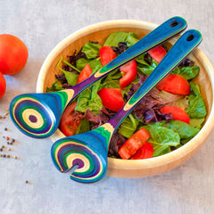 A wooden bowl filled with a fresh salad of mixed greens and sliced tomatoes highlights the vibrant energy of the Baltique® Mumbai Collection Salad Serving Set, 11-3/4" by Totally Bamboo. The colorful, striped serving utensils elegantly rest on top, while two whole tomatoes and scattered peppercorns adorn the gray surface alongside.