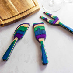 Part of the Baltique® Mumbai Collection from Totally Bamboo, this vibrant 3-piece Cheese Tool Set features cheese knives with striped rainbow handles, artistically arranged on a light surface beside a wooden cutting board. An elegant wine glass stem enhances the charm of this exquisite set.