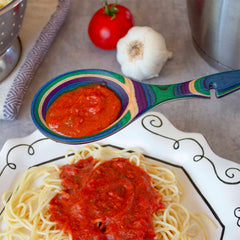 A vibrant Baltique® Mumbai Collection Notched Cooking Spoon by Totally Bamboo holds a serving of tomato sauce above a plate of spaghetti adorned with the same rich sauce. In the background, a fresh tomato, garlic bulb, and colander brimming with pasta complete the scene.