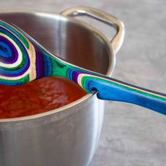 A Baltique® Mumbai Collection Notched Cooking Spoon by Totally Bamboo rests on the edge of a stainless steel pot filled with rich red tomato sauce. This vibrant wooden spoon, crafted from colored birch, features bright swirls of blue, green, purple, and white against a gray textured background.