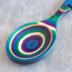 The Baltique® Mumbai Collection Notched Cooking Spoon by Totally Bamboo is a brightly colored birch utensil featuring concentric rings in blue, purple, green, and beige. Set against a textured gray background, this spoon adds a vibrant touch to any kitchen aesthetic.