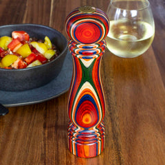 A vibrant striped refillable salt shaker from the Baltique® Marrakesh Collection by Totally Bamboo stands on a wooden table, adding a colorful kick beside a glass of white wine and a bowl of diced tomatoes and yellow peppers.