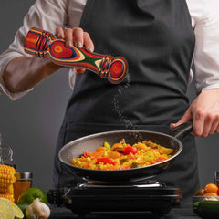 A person in a black apron enhances the vibrant vegetables in the pan with a splash of color. The mixture of peppers, corn, and broccoli is meticulously seasoned using a decorative refillable salt shaker from the Baltique® Marrakesh Collection by Totally Bamboo. Various ingredients are neatly arranged on the countertop.